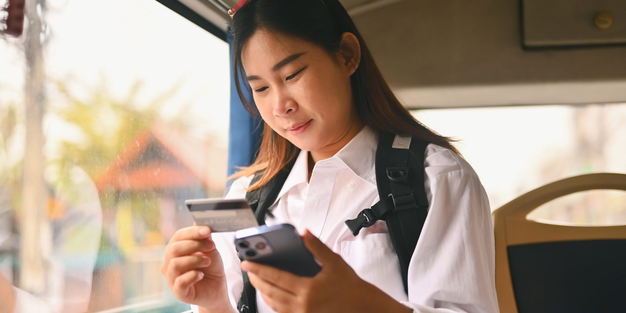 Happy young holding credit card and using mobile phone while sitting inside public bus transport.